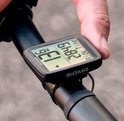 Hand bevestigt zwarte Sigma fietscomputer op fietsstuur. Het scherm toont '13.6 KMH' en '2:8:49 Ride Time'.