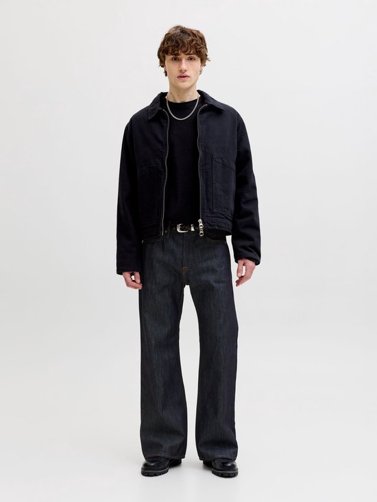Model van voren gezien, draagt een zwarte jas, donkere jeans met wijd uitlopende pijpen en een riem.