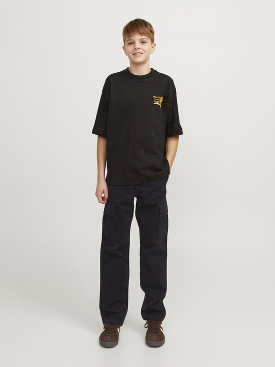 Jongen met een glimlach, draagt een zwart T-shirt met gele print en zwarte cargobroek, staand.