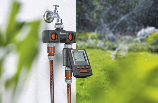 Gardena waterverdeler met kraan en twee slangen. Rechts een digitale timer met scherm. Links een tuin met sproeiers.