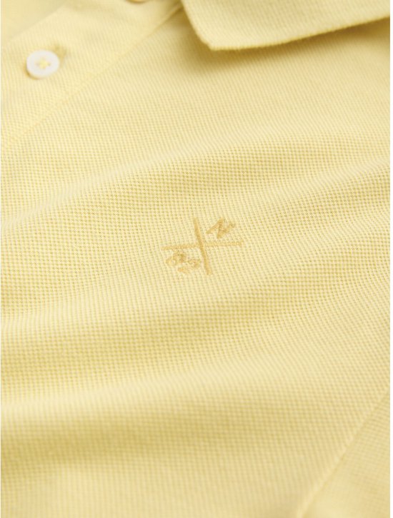 Close-up van het geborduurde logo op een lichtgeel poloshirt.
