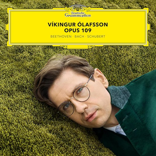 Close-up portret van Vikingur Ólafsson met bril, liggend in groen mos, op de cover van 'Vikingur Ólafsson Opus 109'.