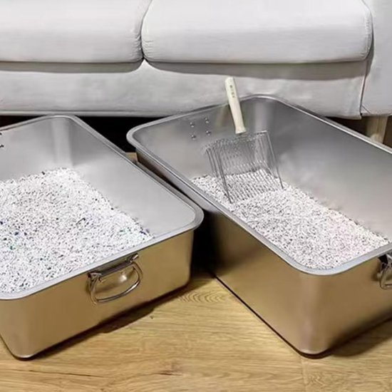 Twee grote zilveren bakken met kattengrit en een schep, geplaatst op een houten vloer bij een witte bank.