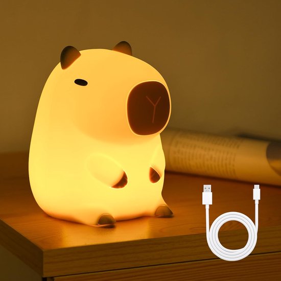 Een geel capibara nachtlampje staat op een houten nachtkastje en gloeit zacht. Een witte USB-kabel is ernaast.