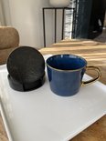 Zwarte ronde onderzetters met marmerprint in een zwarte houder, naast een blauwe mok met gouden handvat op een witte schaal.