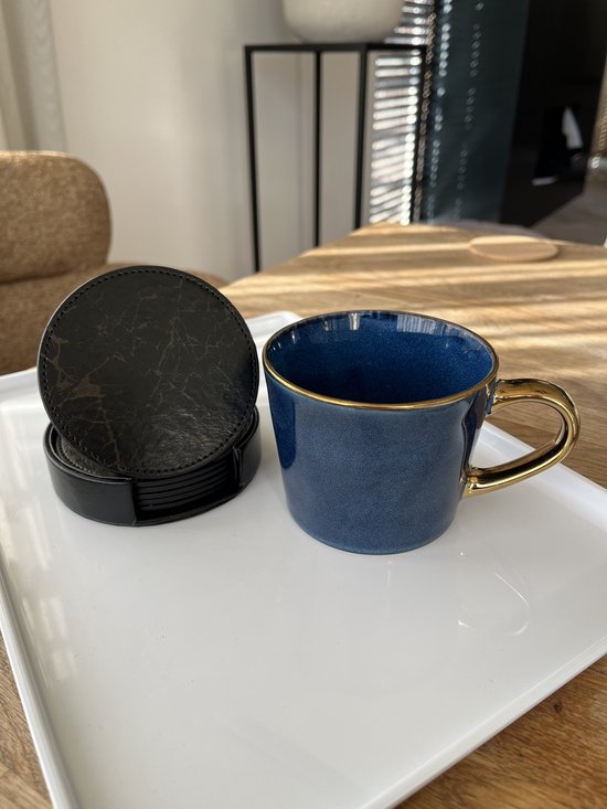 Zwarte ronde onderzetters met marmerprint in een zwarte houder, naast een blauwe mok met gouden handvat op een witte schaal.