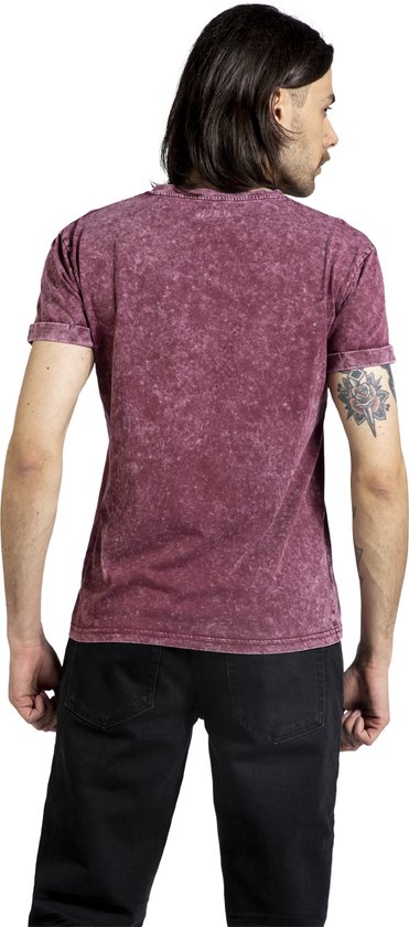 Een man met lang donker haar, in een bordeauxrode, verweerde T-shirt met opgerolde mouwen en zwarte jeans.