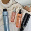 Drie REF-producten: een blauwe textuurspray, een roze shine elixir en een oranje hittebeschermingsspray, naast een krultang.