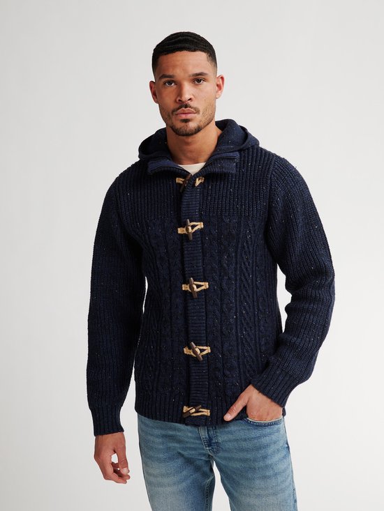 Man in donkerblauwe gebreide kabelvest met capuchon en houten toggle sluitingen, met jeans.