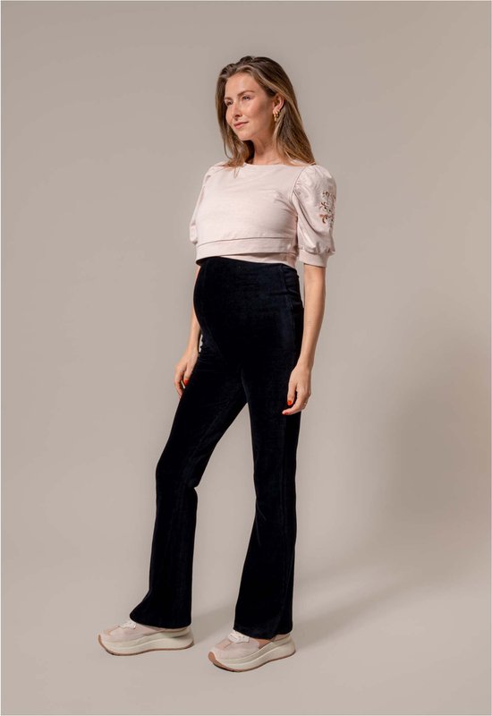 Zijzicht van een zwangere vrouw in een donkerblauwe, uitlopende corduroy zwangerschapsbroek en beige crop top met borduursels op de mouwen.