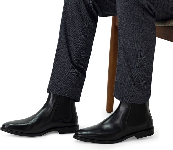 Un homme en pantalon bleu foncé, assis, portant des chelsea boots noires et brillantes.