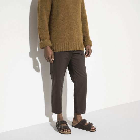Man draagt donkerbruine Birkenstock Arizona pantoffels met lammenvacht voering, gecombineerd met een bruine trui en donkere broek.