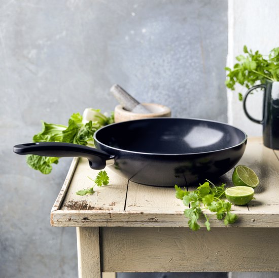 Zwarte wokpan met steel, limoenpartjes en koriander op een rustieke tafel.