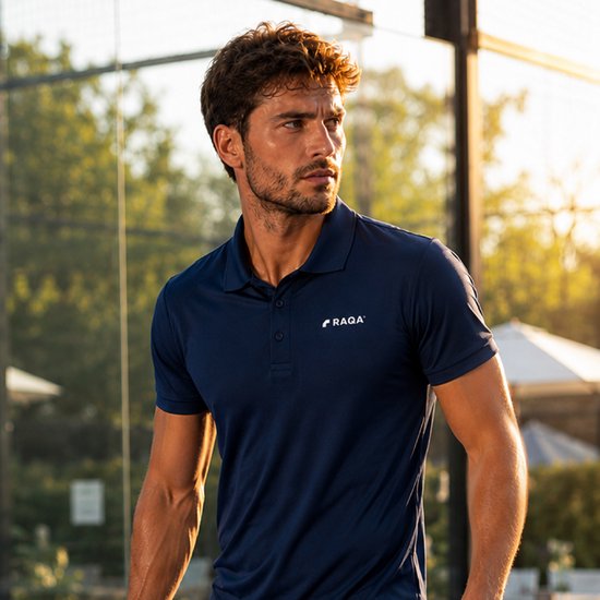 Man draagt een donkerblauwe sportpolo met korte mouwen en het Raqa-logo op de borst. Hij staat buiten in de zon.