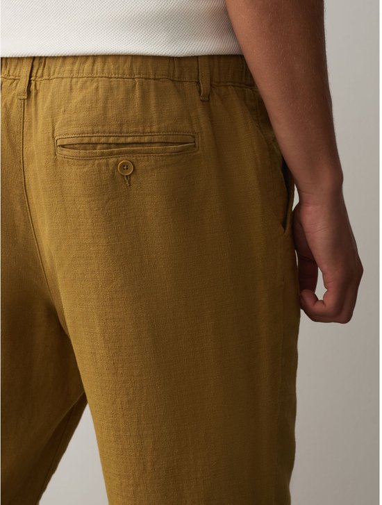 Close-up van de achterkant van een okergele broek van linnenmix, met focus op de paspelzak met knoop.