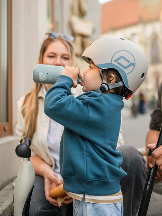 Kind draagt een crèmekleurige helm met blauwe logo's en drinkt uit een flesje.