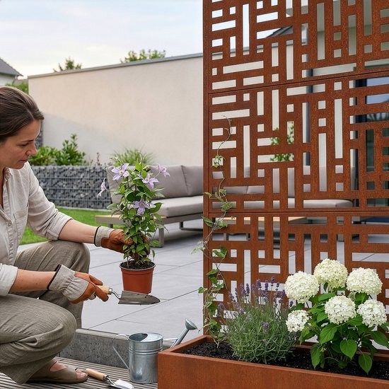 Vrouw met tuinhandschoenen plant bloemen in roestkleurige plantenbak naast tuinscherm.