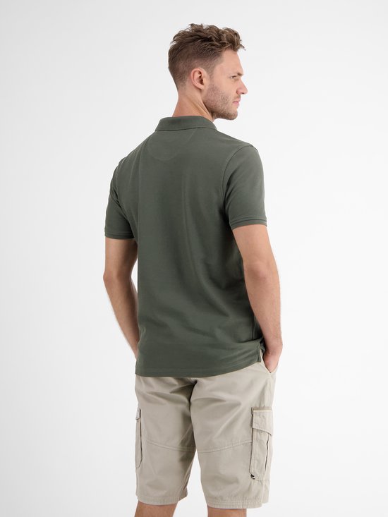 Man met donkergroen poloshirt en beige cargo shorts, staand met handen in de zakken.