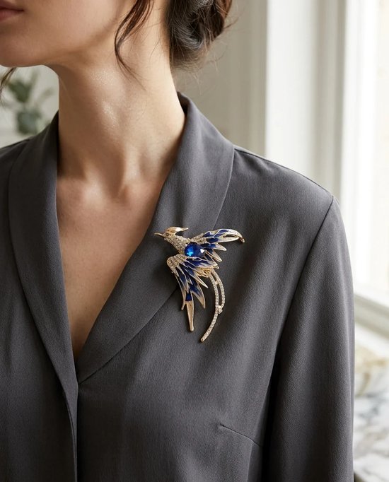 Vrouw draagt een gouden broche met blauwe en witte kristallen in de vorm van een vogel op een grijs jasje.