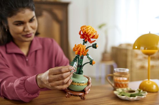 Une femme assise à une table construit un chrysanthème Lego. Les fleurs sont orange et rouges.