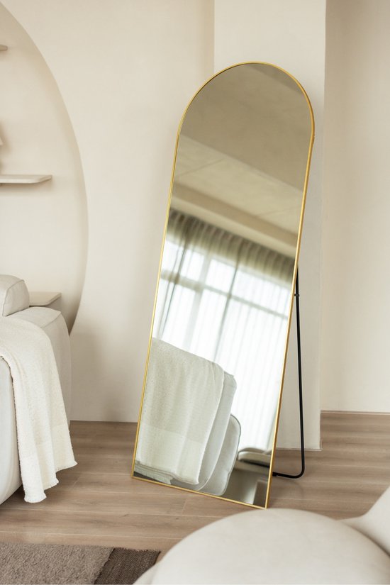Hoge, smalle gouden spiegel met afgeronde bovenkant, reflecteert witte handdoeken, in een lichte woonkamer.