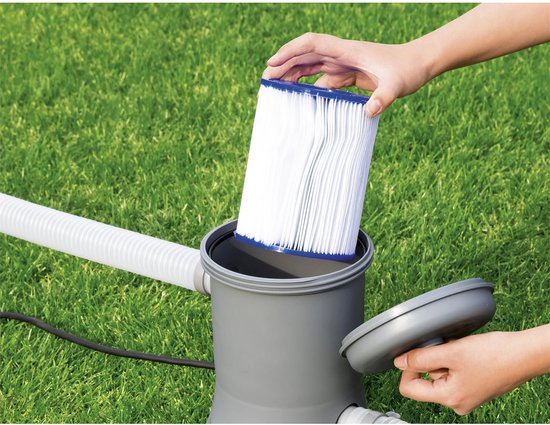 Een persoon vervangt een wit pliss filter met blauwe uiteinden in een grijze zwembadpomp op groen gras.