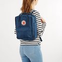 Vrouw met marineblauwe Kånken rugzak op haar rug, draagt een gestreept shirt en spijkerbroek.