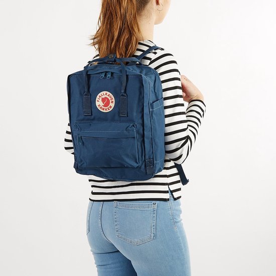 Vrouw met marineblauwe Kånken rugzak op haar rug, draagt een gestreept shirt en spijkerbroek.
