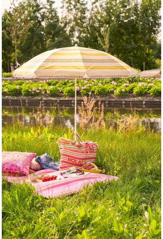 Strandparasol met gele en witte strepen, met een tas met het woord 'HOLA', kussens en picknickspullen op een deken in het gras.