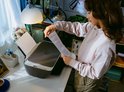 Vrouw in beige trui plaatst een geprint vel papier in een zwarte HP DeskJet printer op een wit bureau.
