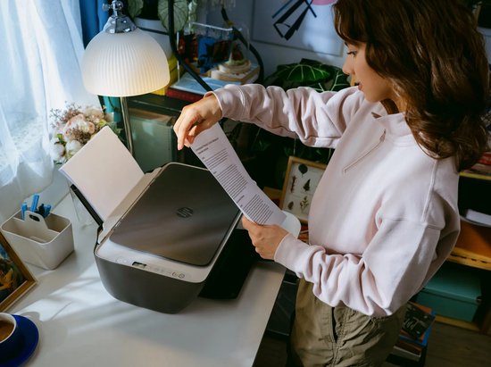 Vrouw in beige trui plaatst een geprint vel papier in een zwarte HP DeskJet printer op een wit bureau.