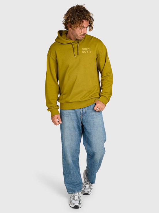Staande man in een olijfgroene Brunotti hoodie en wijde blauwe jeans, volledige lengte.