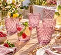 Quatre verres roses à motif nid d'abeille sur une table en bois avec des fruits et des serviettes.