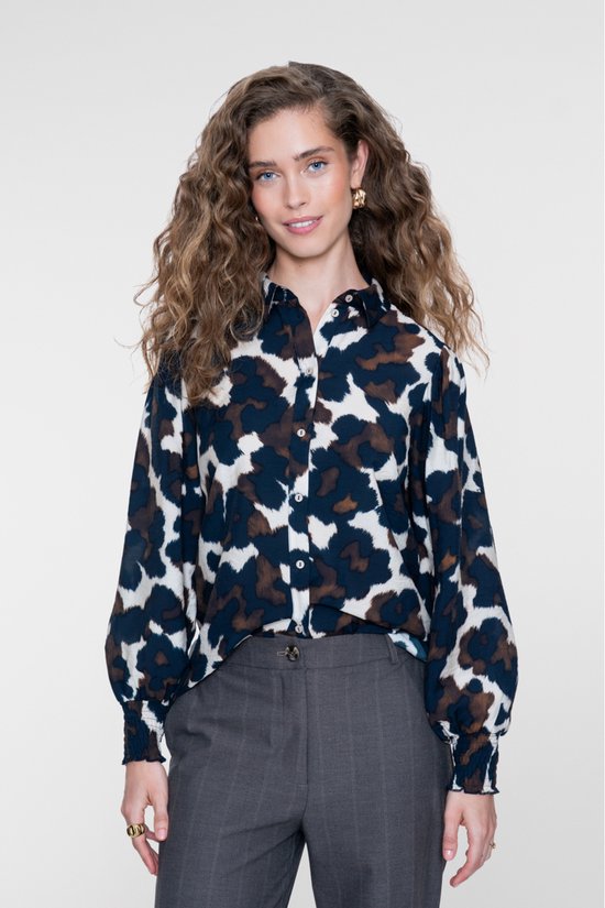 Model draagt een blouse met abstracte print in blauw, bruin en crème, gecombineerd met een grijze pantalon.