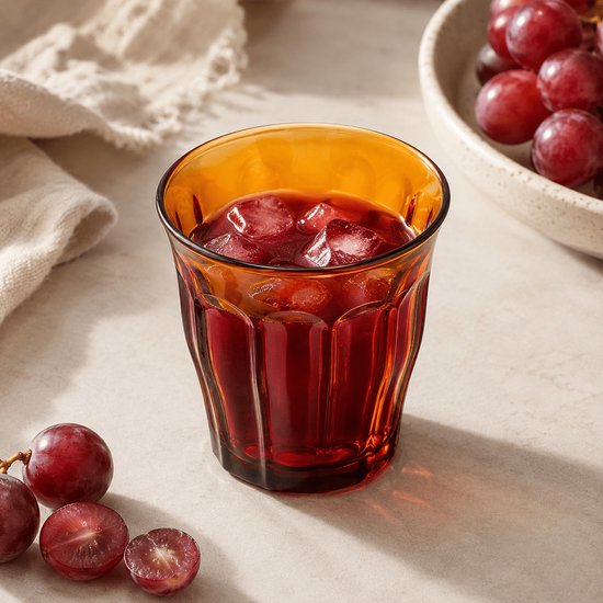 Un verre Duralex Picardie ambré avec des glaçons et une boisson rouge, à côté de raisins.