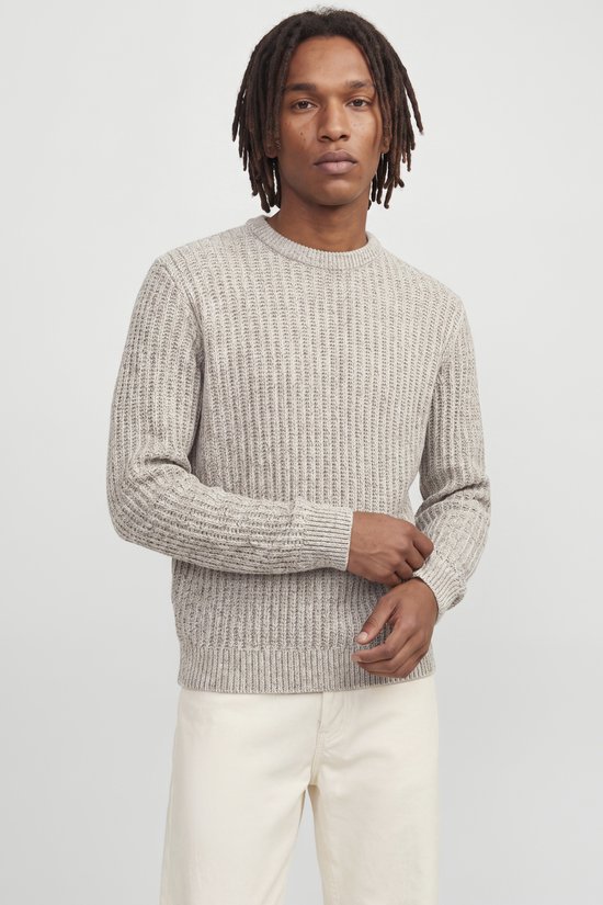 Model met dreadlocks in beige trui en witte broek, kijkt naar de camera.