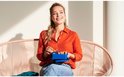 Femme en chemise orange et jean, assise, tenant une boîte à lunch bleue avec des amuse-gueules.