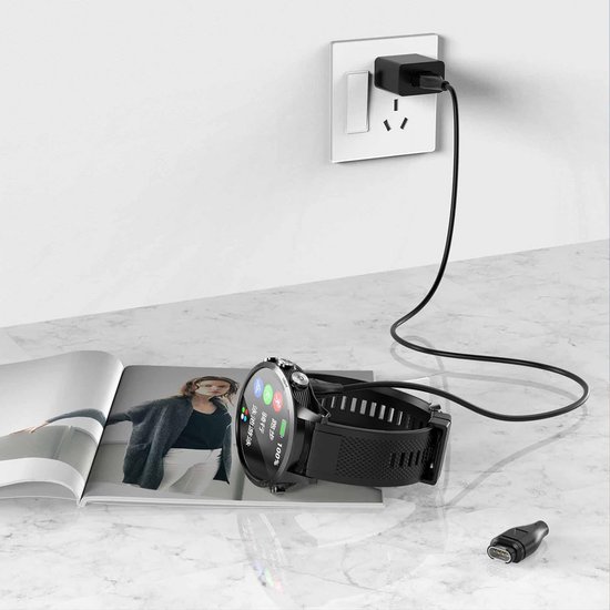 Zwarte smartwatch met oplaadkabel aangesloten op een stopcontact, op een tijdschrift.