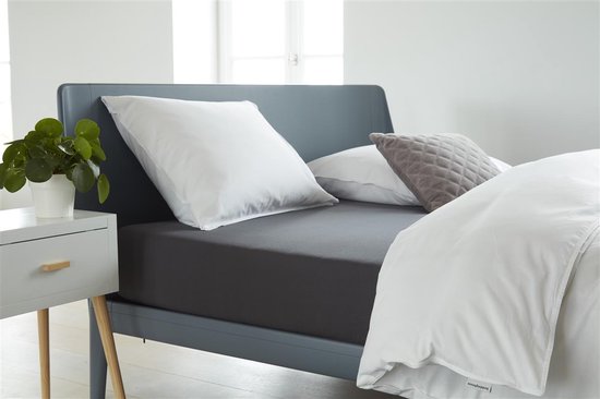 Een bed met een donkergrijs jersey topper hoeslaken, witte kussens en dekbed, en een plant.