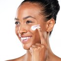Vrolijke donkere vrouw met glimlach brengt witte crème aan op haar wang met haar vinger.