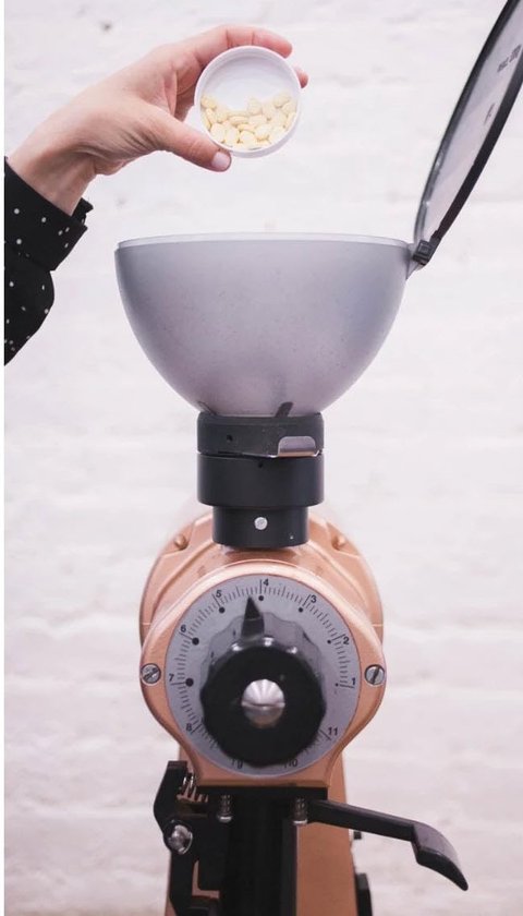 Een hand leegt Urnex Grindz reinigingstabletten uit een pot in de hopper van een koperkleurige koffiemolen.