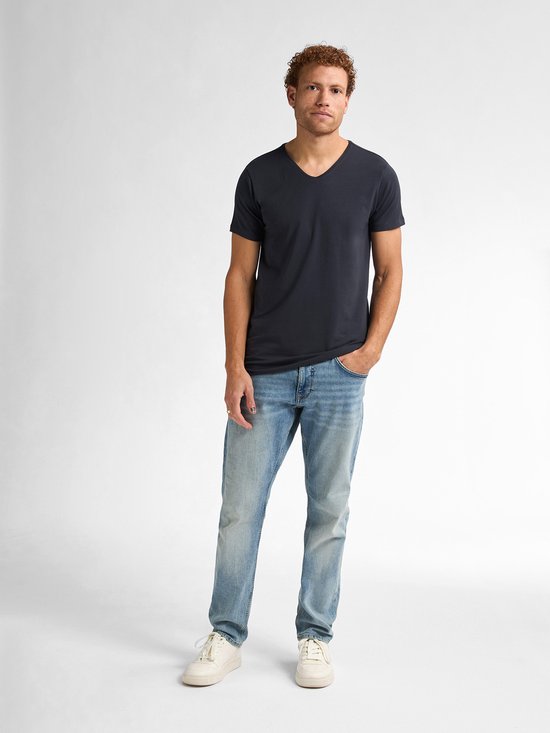 Man met krullend bruin haar draagt een donkerblauwe V-hals T-shirt, spijkerbroek en witte sneakers.