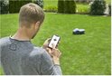 Man in grijs shirt bedient robotmaaier op gazon via smartphone.