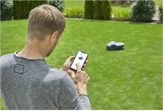 Man in grijs shirt bedient robotmaaier op gazon via smartphone.