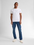 Man staat frontaal in een witte t-shirt met logo en een blauwe spijkerbroek, rechte pasvorm, met witte sneakers.