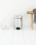 Briljant stalen Brabantia NewIcon prullenbak met zwart voetpedaal, naast een houten bankje met handdoek.