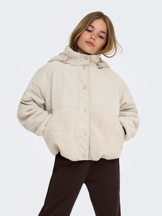 Meisje staat met handen in de zakken, draagt een beige teddy jas, bruine joggingbroek.
