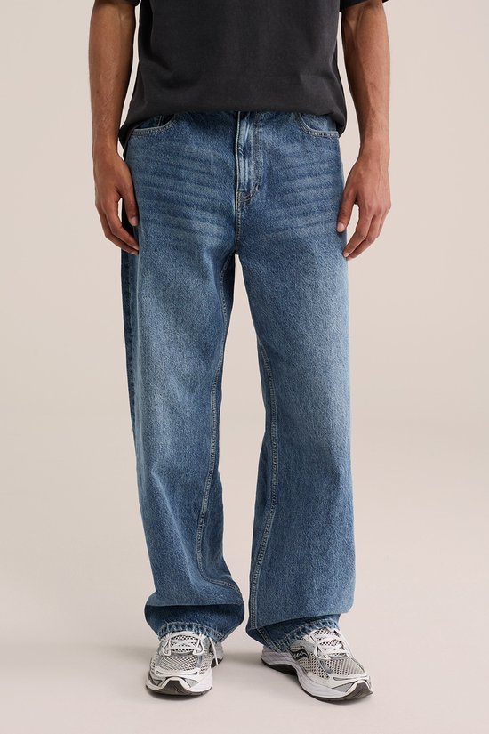 Voorkant van een man in een loszittende, blauwe spijkerbroek en grijze sneakers. De broek heeft een ritssluiting en knoop.