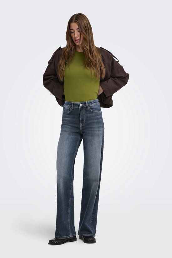 Model in een olijfgroene coltrui, bruine jas en wijde spijkerbroek met een stonewash-look.