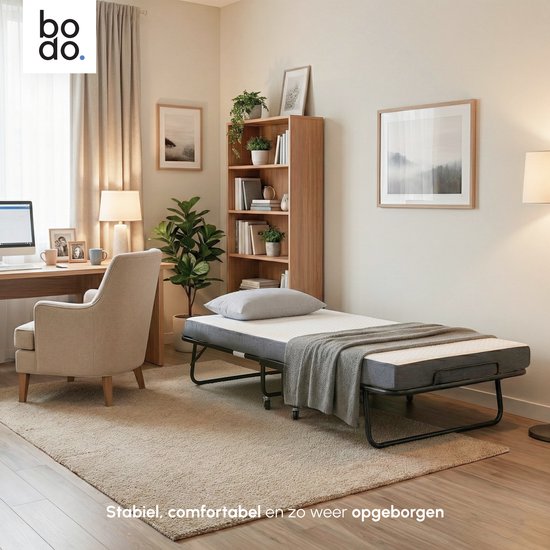 Uitgeklapt Bodo logeerbed in een woonkamer, met een vrouw die leest. Het bed heeft een donkergrijs matras, een zwart frame en wieltjes.